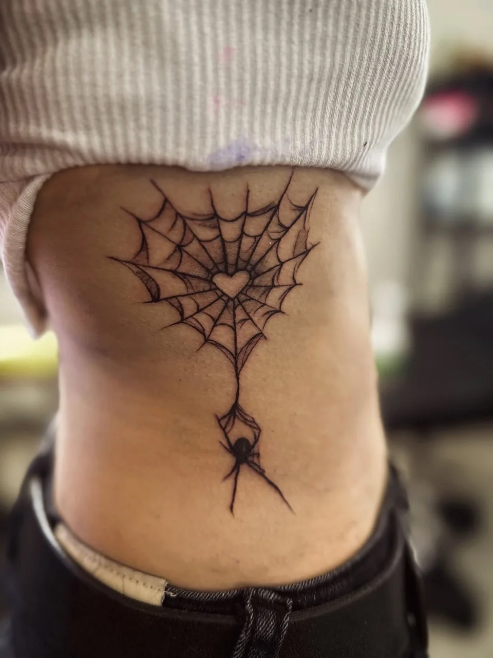 Tatuaż pajęczyny z serduszkiem i zwisającym pająkiem na żebrach, czarny tusz – SHADY TATTOO Wieluń