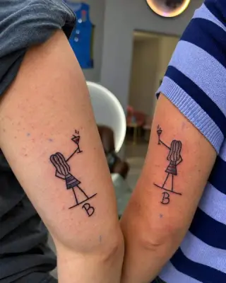 Dwa takie same tatuaże na tylnej stronie ramion dwóch osób: kreskowa postać z kieliszkiem i literą B, czarna linia – matching tattoo z SHADY TATTOO w Wieluniu