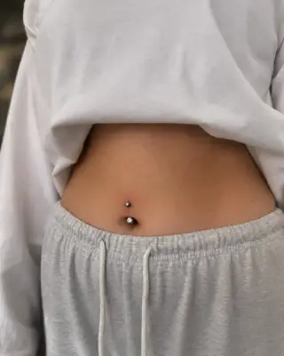 Piercing pępka z kolczykiem z cyrkonią, klientka w szarych spodniach dresowych