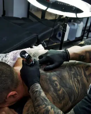 Tatuator przy pracy w studiu w Wieluniu, wykonuje rozległy tatuaż na plecach klienta pod lampą pierścieniową
