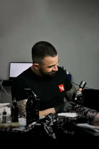 Brodaty tatuator w czarnej koszulce przy pracy maszynką w studiu SHADY TATTOO, ujęcie z boku